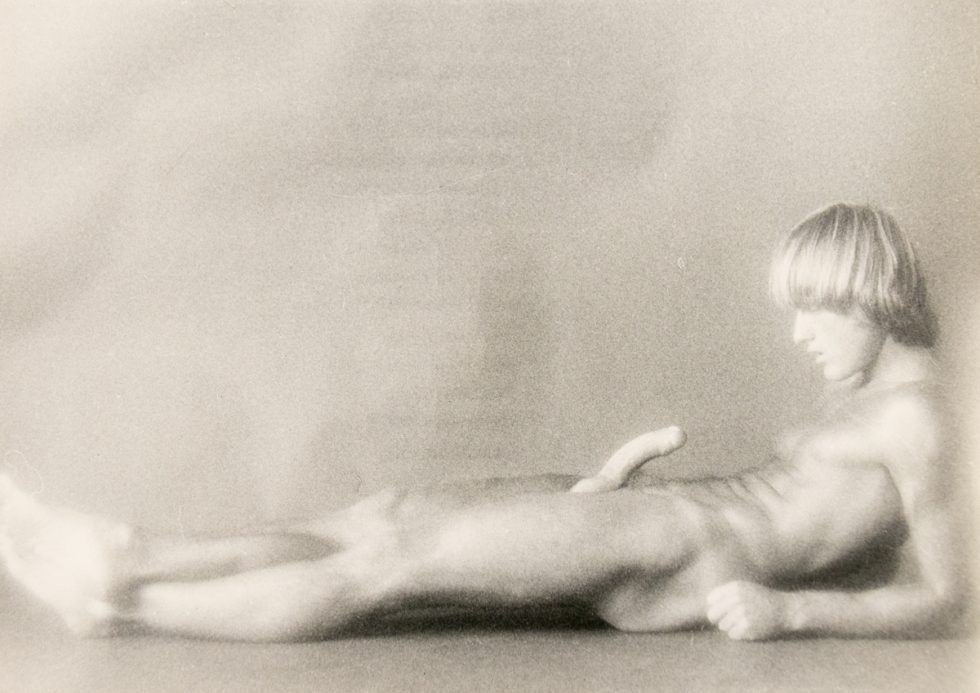 A nude figure reclines on the floor, gazing downward, in a softly lit and blurred black-and-white composition.