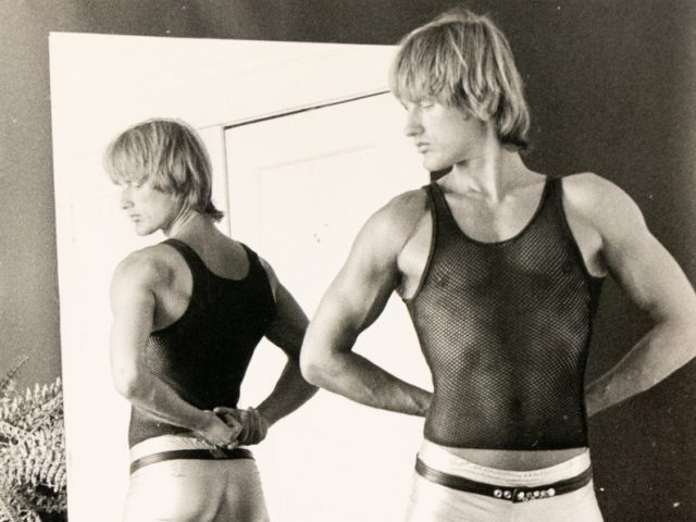 A black and white photograph of a man in a black tank top and silver metallic pants.
