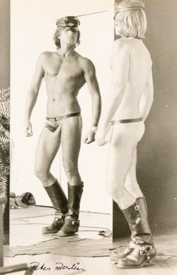 A shirtless man wearing a cap, a thong, and boots looks at himself in a mirror, revealing his reflection from behind.