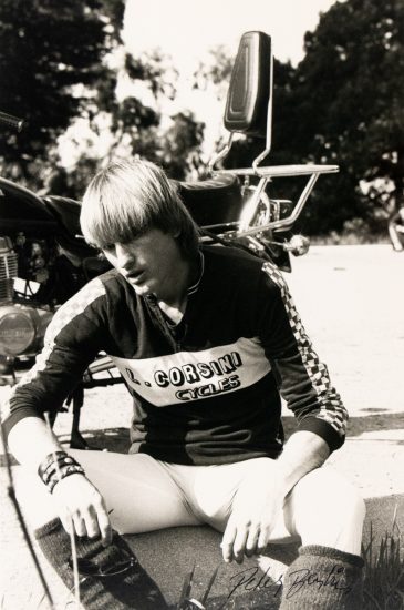 A black and white portrait of a man with a bowl cut sitting by a motorcycle.