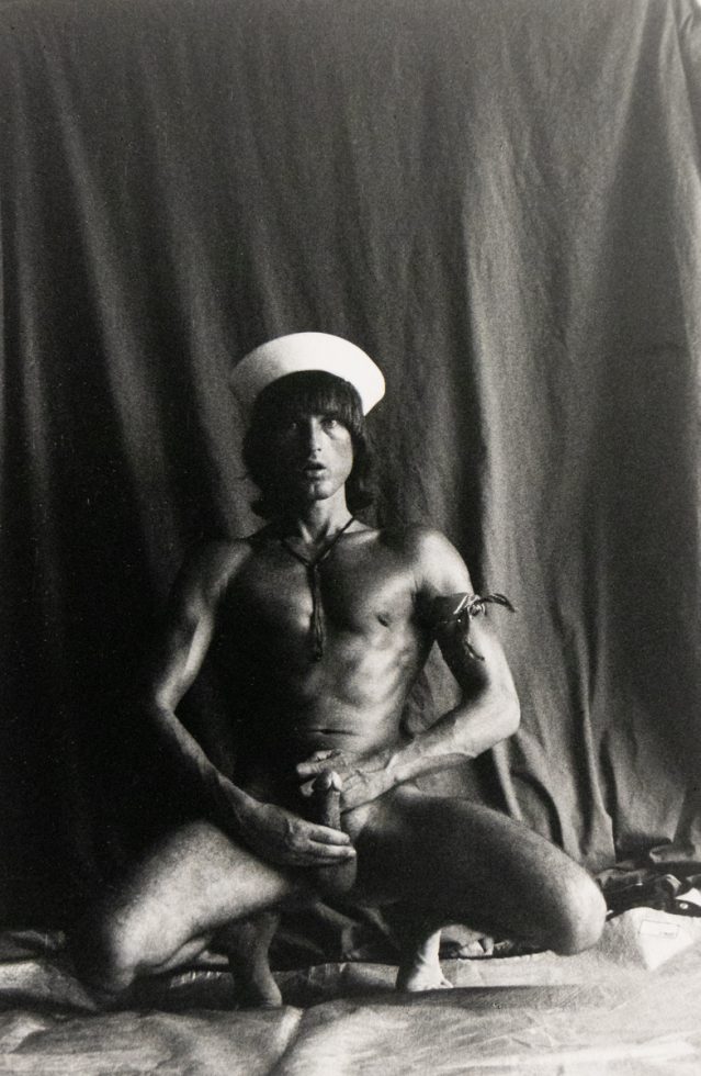 A black and white photograph of a nude man wearing a sailor hat.