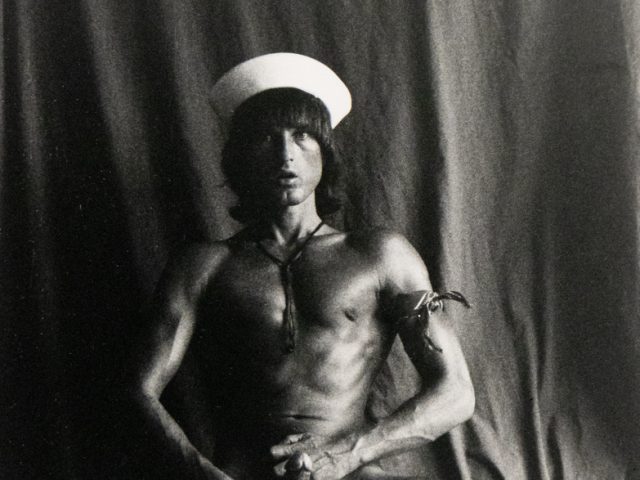A black and white photograph of a nude man wearing a sailor hat.