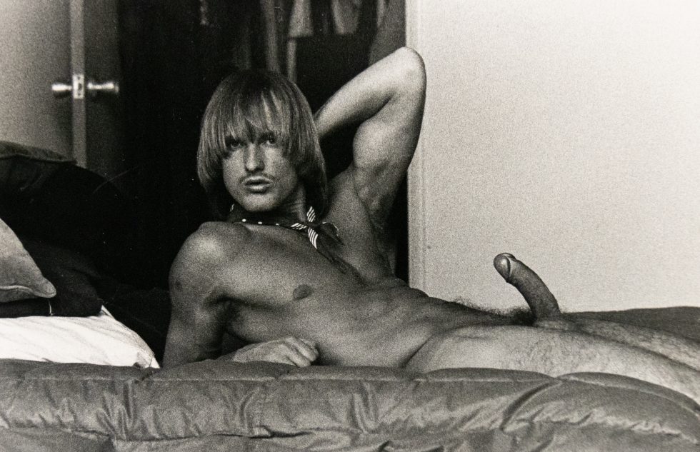 A black and white photograph of a nude man with a bowl cut reclining on a bed with an erection.