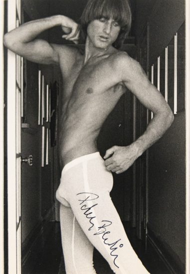 Shirtless person in white tights poses in a hallway, flexing an arm and turning slightly; autograph appears on the thigh.