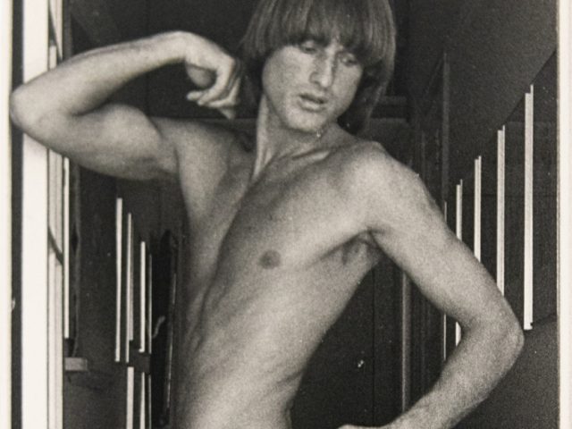 Shirtless person in white tights poses in a hallway, flexing an arm and turning slightly; autograph appears on the thigh.