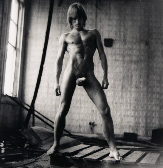 Nude man standing with legs apart on wooden boards in a dilapidated room, lit from a window on the left.