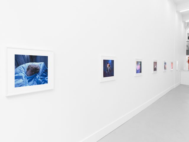 Brightly lit gallery with colorful, surreal portraits in white frames displayed along stark white walls.