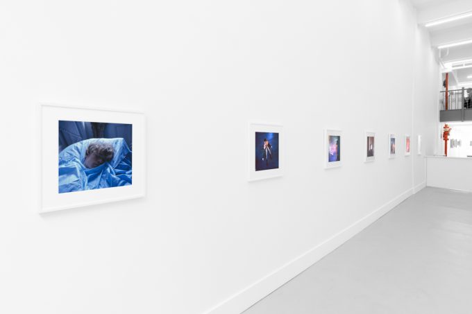 Brightly lit gallery with colorful, surreal portraits in white frames displayed along stark white walls.
