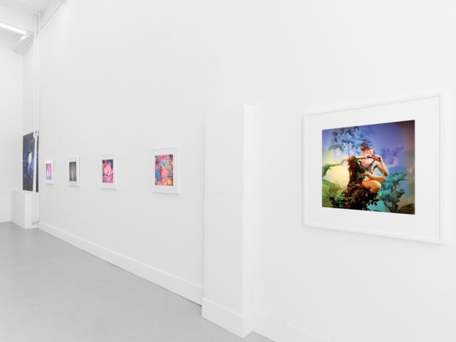 Brightly lit gallery with colorful, surreal portraits in white frames displayed along stark white walls.