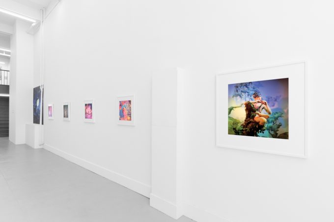 Brightly lit gallery with colorful, surreal portraits in white frames displayed along stark white walls.
