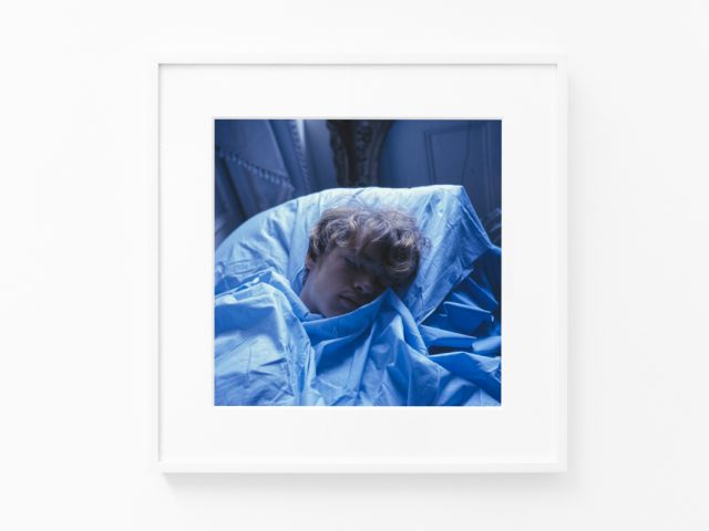 Brightly lit gallery with colorful, surreal portraits in white frames displayed along stark white walls.