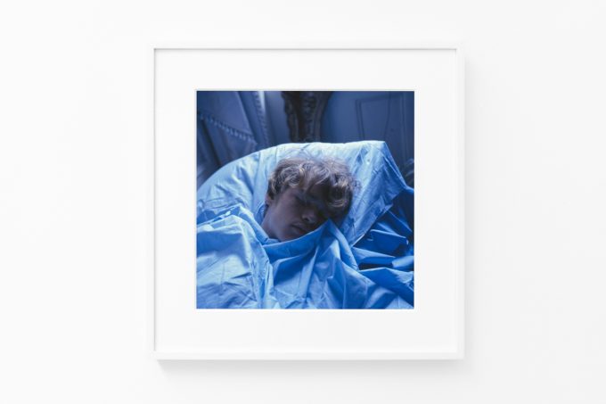 Brightly lit gallery with colorful, surreal portraits in white frames displayed along stark white walls.