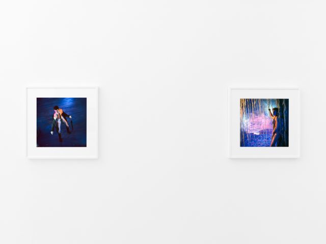 Brightly lit gallery with colorful, surreal portraits in white frames displayed along stark white walls.
