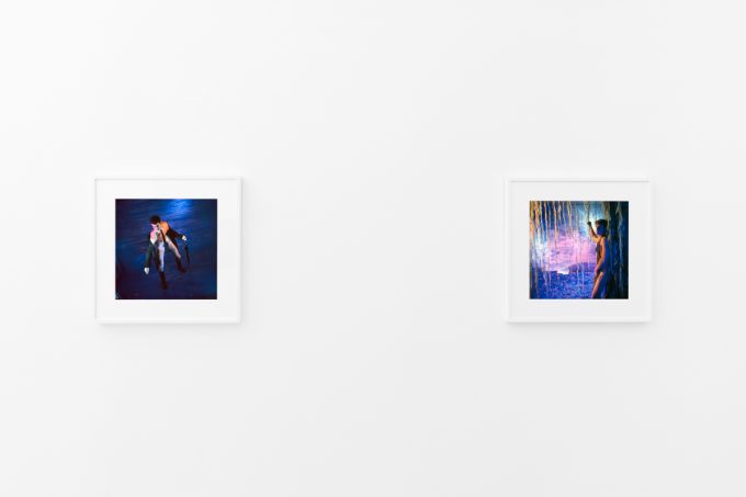 Brightly lit gallery with colorful, surreal portraits in white frames displayed along stark white walls.