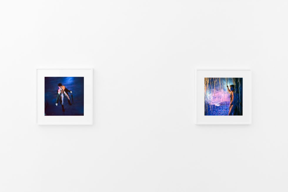 Brightly lit gallery with colorful, surreal portraits in white frames displayed along stark white walls.