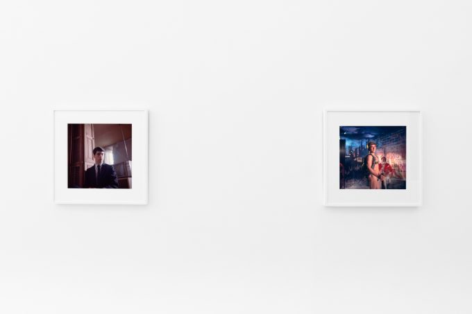 Brightly lit gallery with colorful, surreal portraits in white frames displayed along stark white walls.