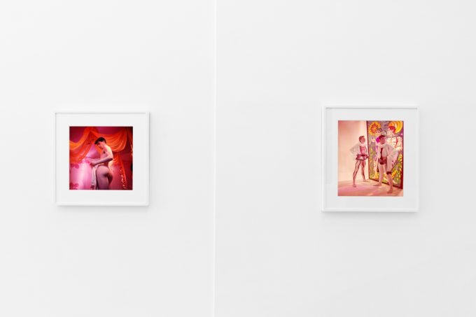 Brightly lit gallery with colorful, surreal portraits in white frames displayed along stark white walls.