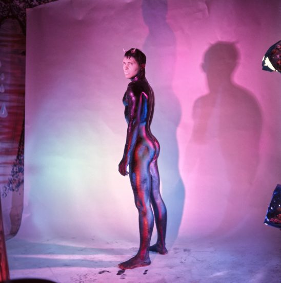 A slender, body-painted person with small horns stands facing left, looking back at the viewer, against a pink and blue lit wall.