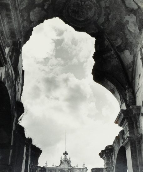 Crumbled stone arches frame a cloudy sky above a distant ornate facade with a cross-topped spire.