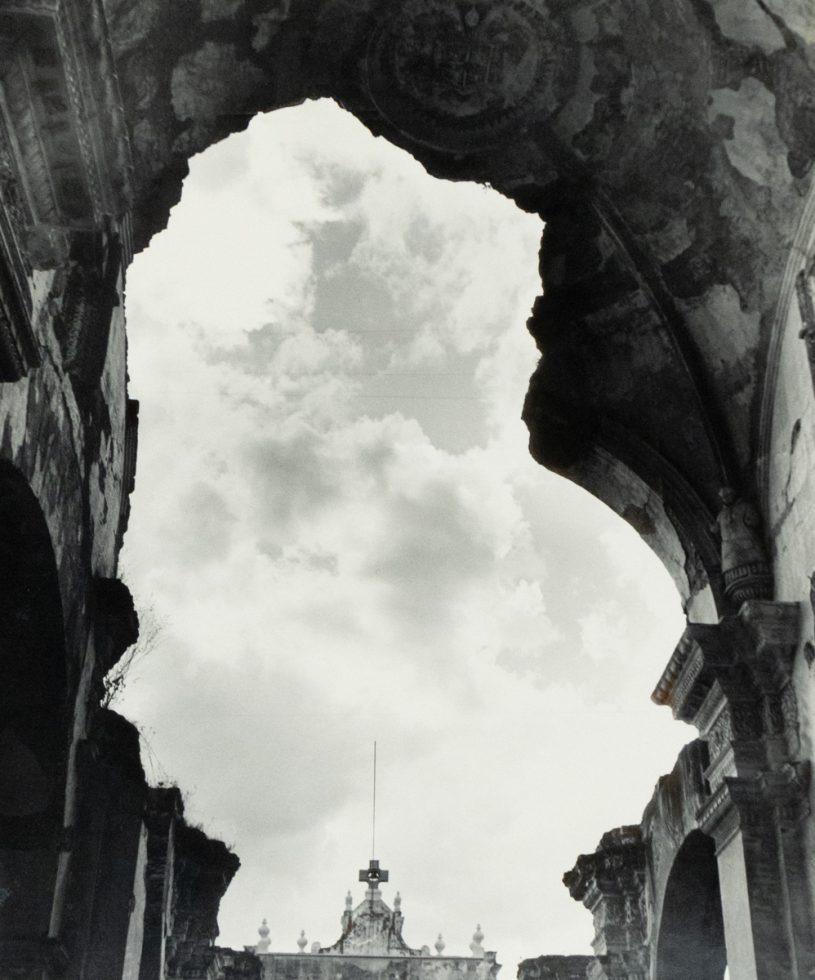 Crumbled stone arches frame a cloudy sky above a distant ornate facade with a cross-topped spire.