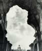 Crumbled stone arches frame a cloudy sky above a distant ornate facade with a cross-topped spire.