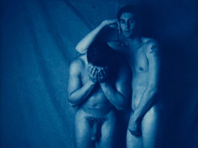 Two nude figures stand close; one covers his face while the other rests a hand gently on his head.