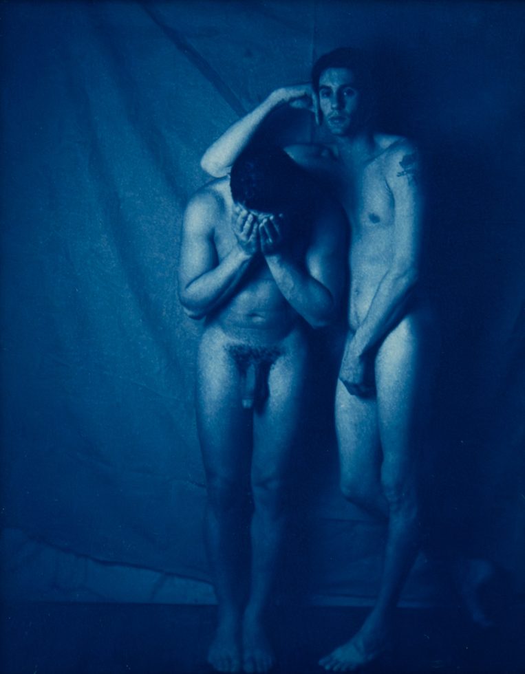 Two nude figures stand close; one covers his face while the other rests a hand gently on his head.