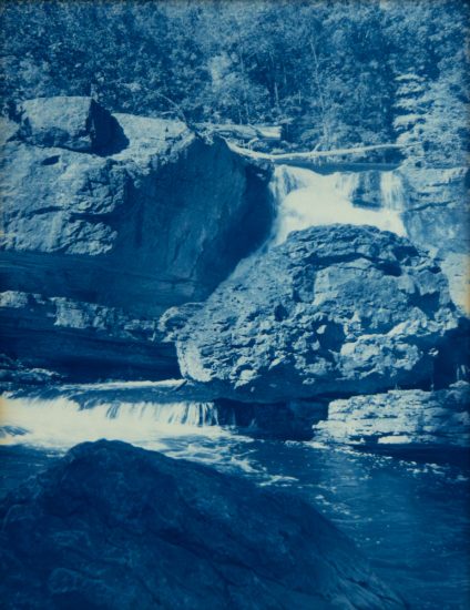 Water cascades over layered rock formations into a calm pool, surrounded by dense trees in a deep blue cyanotype print.