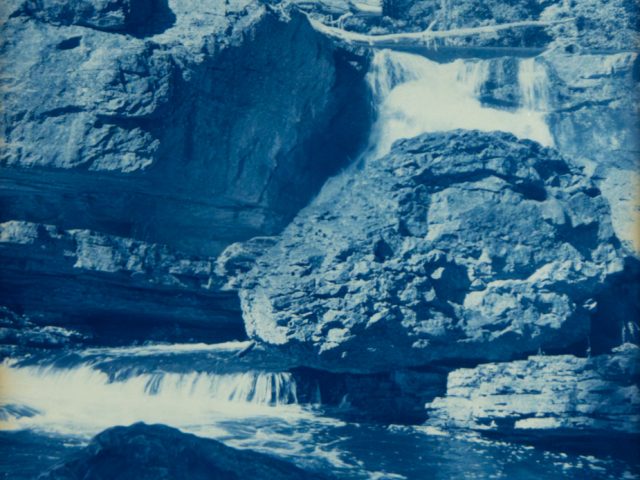 Water cascades over layered rock formations into a calm pool, surrounded by dense trees in a deep blue cyanotype print.