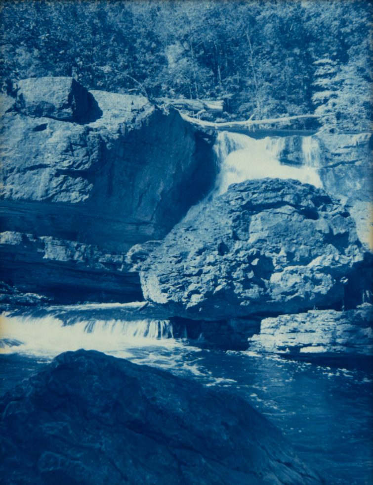 Water cascades over layered rock formations into a calm pool, surrounded by dense trees in a deep blue cyanotype print.