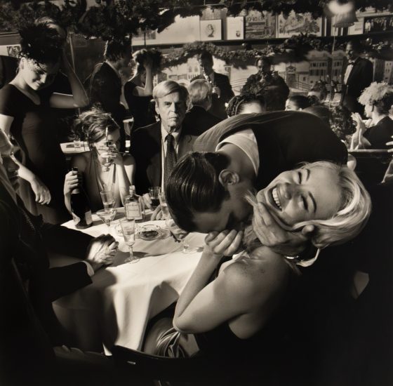 A black and white photograph captures a lively and somewhat chaotic scene in a restaurant, with people interacting closely around a table, and one man affectionately kissing a woman's neck while she laughs.