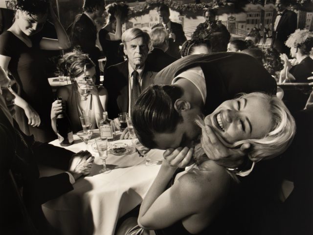 A black and white photograph captures a lively and somewhat chaotic scene in a restaurant, with people interacting closely around a table, and one man affectionately kissing a woman's neck while she laughs.