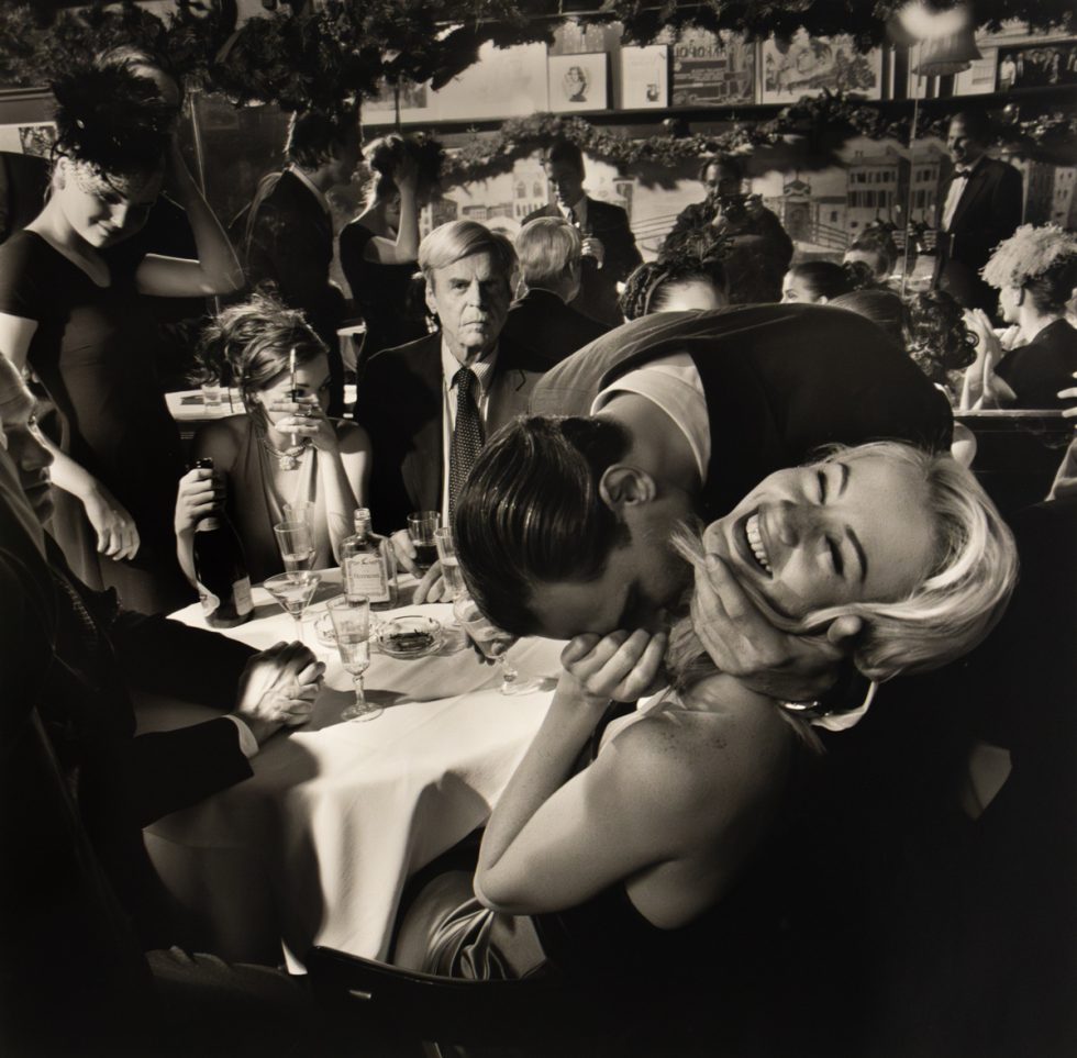 A black and white photograph captures a lively and somewhat chaotic scene in a restaurant, with people interacting closely around a table, and one man affectionately kissing a woman's neck while she laughs.