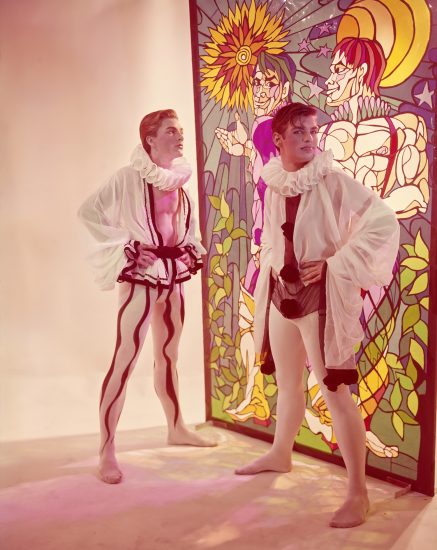 Click for sizes / editions / prices. Two pale-skinned performers in white ruffled collars and harlequin-style costumes stand before a colorful stained-glass panel depicting figures.