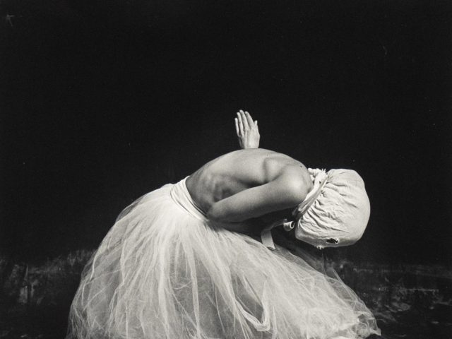 A lone figure, draped in white, kneels with bowed head and upraised hands against a dark void, evoking a sense of sorrow or profound contemplation.
