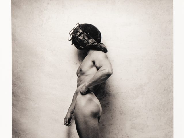 A profile shot of a person with a fit physique, wearing a protective headpiece with a face covering, against a textured background.