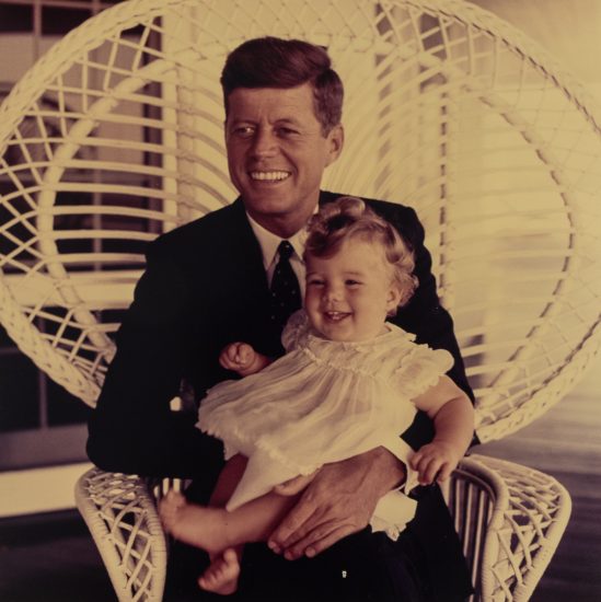 A color photograph of JFK sitting in a white wicker chair golding his smiling daughter.