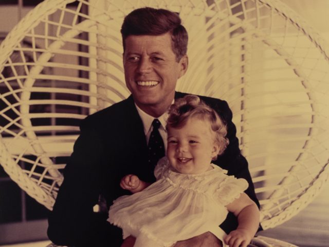 A color photograph of JFK sitting in a white wicker chair golding his smiling daughter.