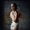 A cinematic portrait of an adolescent girl with red hair and freckles, wearing a white undergarment against a dark, textured wall.
