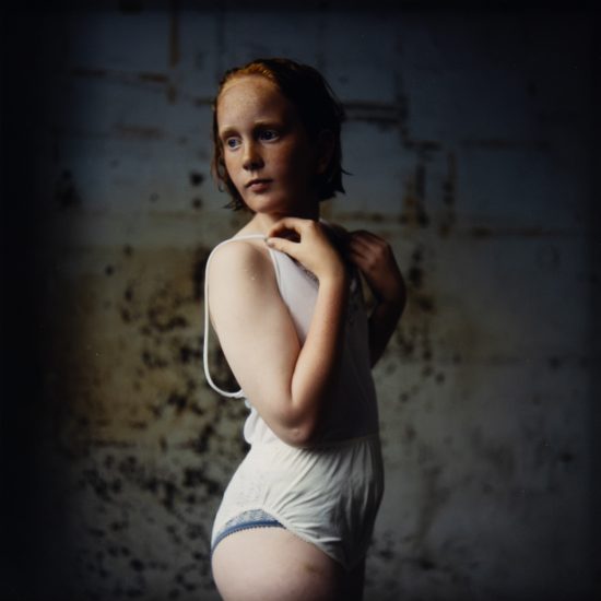 A cinematic portrait of an adolescent girl with red hair and freckles, wearing a white undergarment against a dark, textured wall.