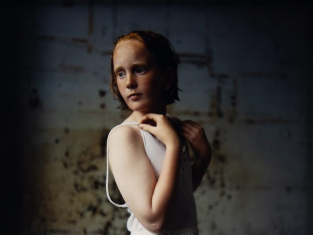 A cinematic portrait of an adolescent girl with red hair and freckles, wearing a white undergarment against a dark, textured wall.