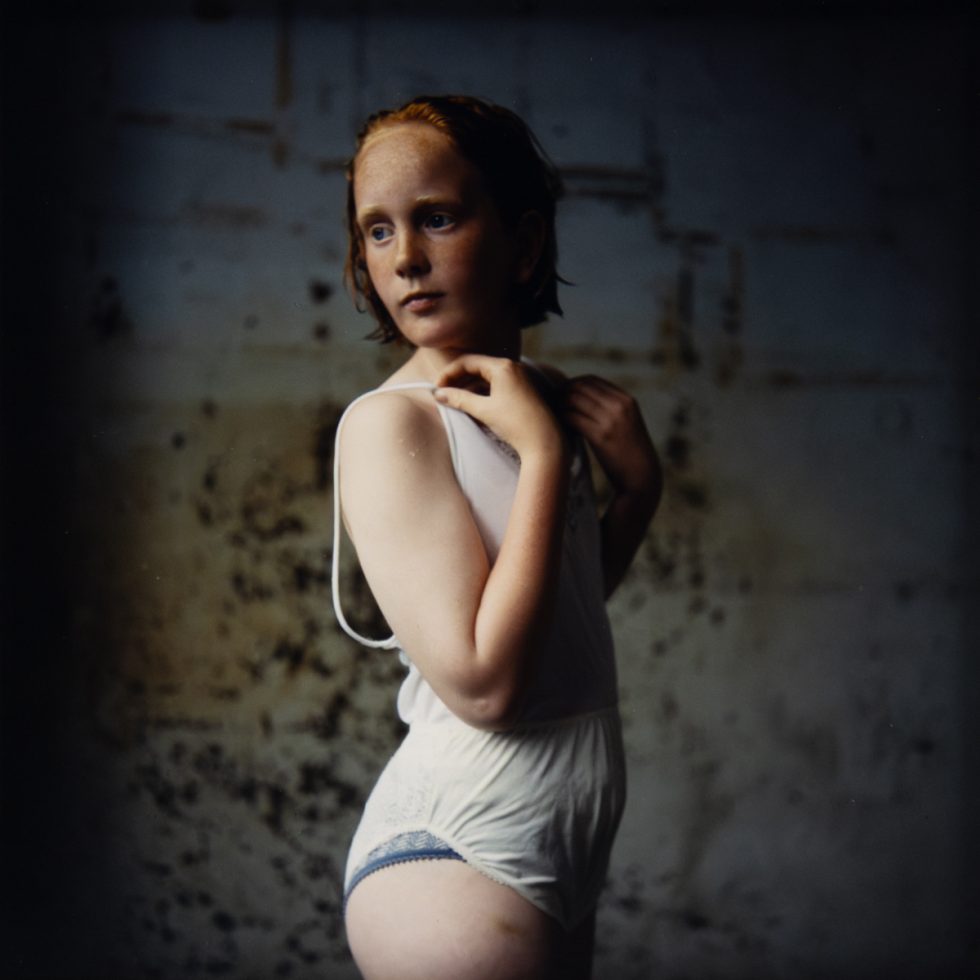 A cinematic portrait of an adolescent girl with red hair and freckles, wearing a white undergarment against a dark, textured wall.
