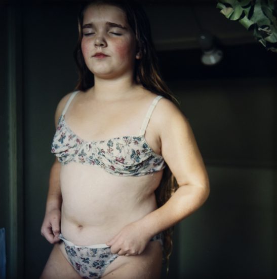 A young girl with long hair stands indoors wearing floral-patterned undergarments, eyes closed and expression calm.