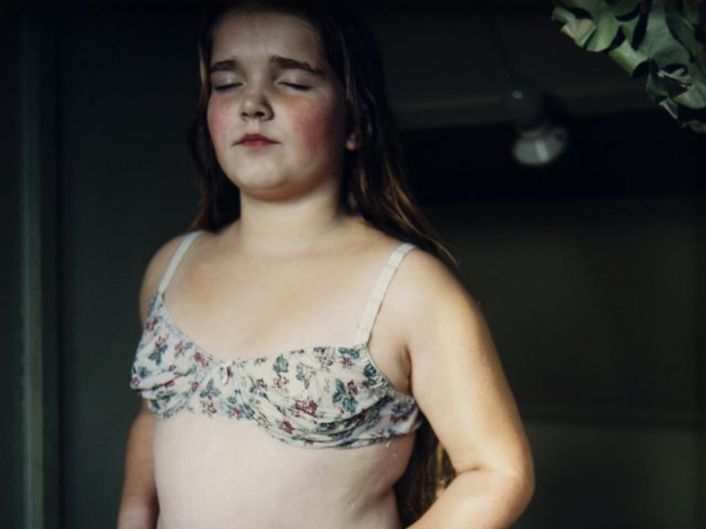 A young girl with long hair stands indoors wearing floral-patterned undergarments, eyes closed and expression calm.