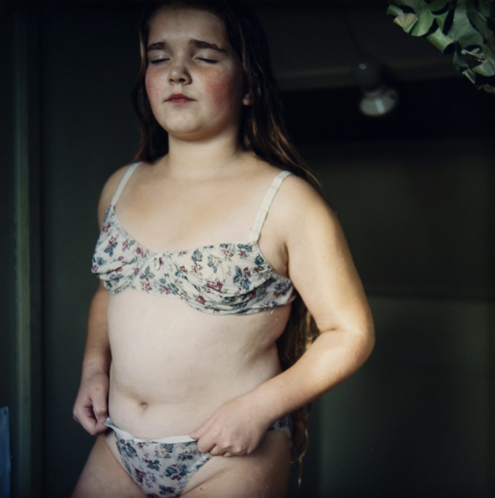 A young girl with long hair stands indoors wearing floral-patterned undergarments, eyes closed and expression calm.