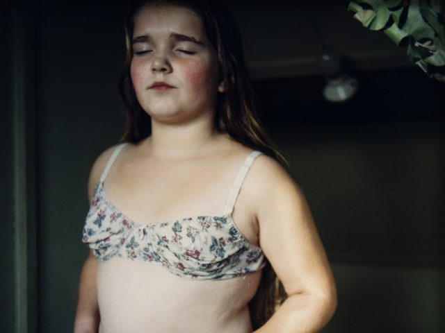 A young girl with long hair stands indoors wearing floral-patterned undergarments, eyes closed and expression calm.