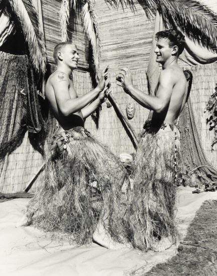 Two young men in grass skirts face each other playfully, mid-dance, in a tropical-themed setting with palm fronds and bamboo walls.
