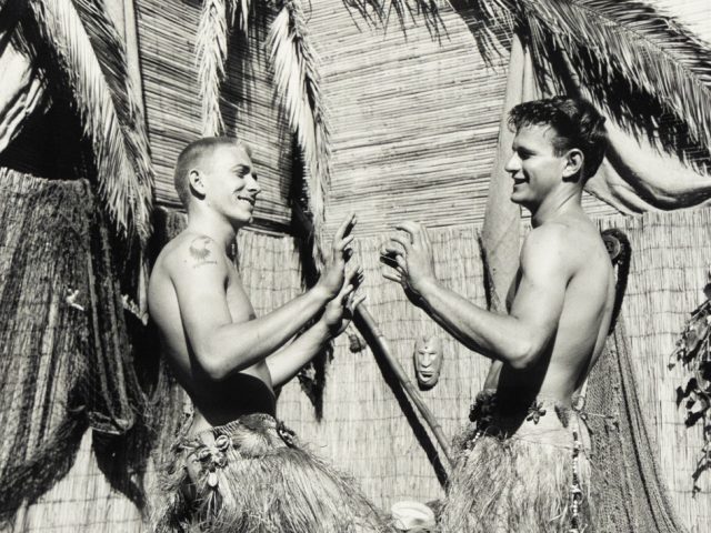 Two young men in grass skirts face each other playfully, mid-dance, in a tropical-themed setting with palm fronds and bamboo walls.