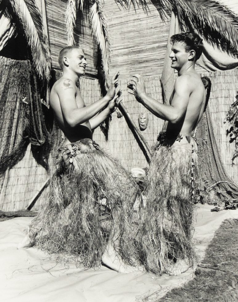 Two young men in grass skirts face each other playfully, mid-dance, in a tropical-themed setting with palm fronds and bamboo walls.