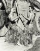 Two young men in grass skirts face each other playfully, mid-dance, in a tropical-themed setting with palm fronds and bamboo walls.